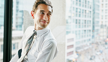 A young businessman sits in an office overlooking a city while engaging in a casual conversation with someone out of view.