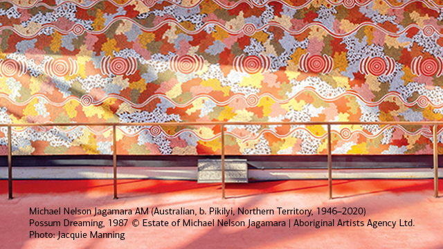 A mural by Michael Nelson Jagamara features layered Aboriginal patterns in warm reds, oranges, yellows, and white.