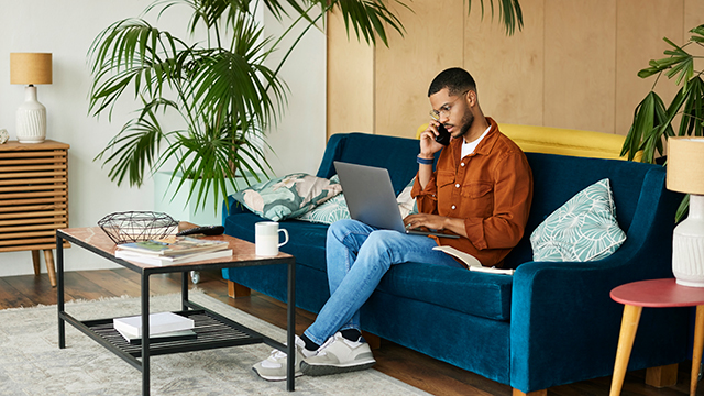 Man on couch working on computer while talking on phone